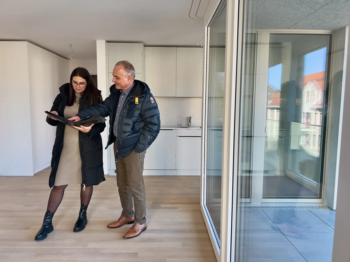 Zwei Menschen stehen vor einer Küche in einem Wohnraum und schauen gemeinsam auf Dokumente in einer Arbeitsmappe.